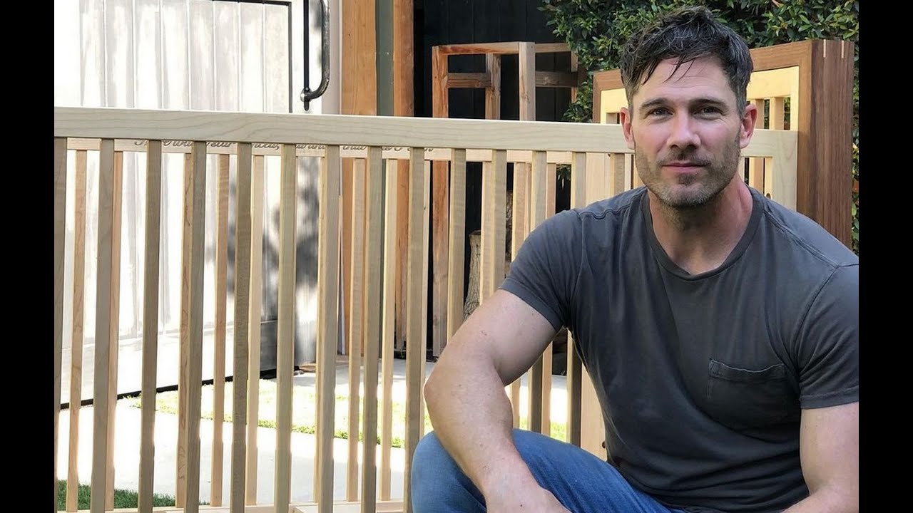 A man in a gray t-shirt kneels next to a wooden crib outdoors, with wooden structures in the background.