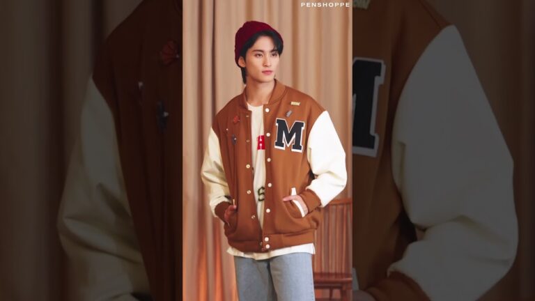 A person wearing a brown letterman jacket, white shirt, jeans, and red beanie, standing indoors.