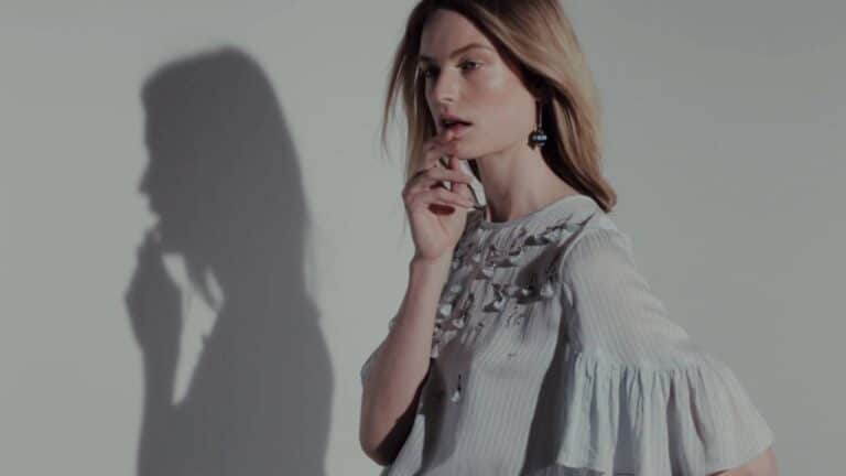 Woman with long hair posing in a light-colored blouse with embroidered patterns, resting one finger on her chin; her shadow is visible on the wall behind her.