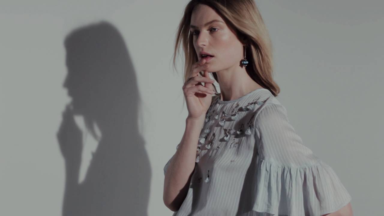 Woman with long hair posing in a light-colored blouse with embroidered patterns, resting one finger on her chin; her shadow is visible on the wall behind her.