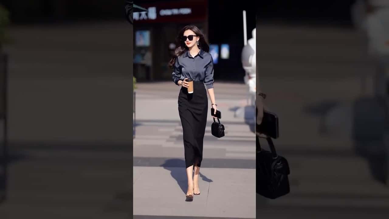 A woman in sunglasses walks outdoors, wearing a dark long-sleeve shirt, black skirt, and shoes. She carries a small black bag and a cup.