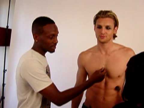 Person standing shirtless while another adjusts clothing in a studio setting.