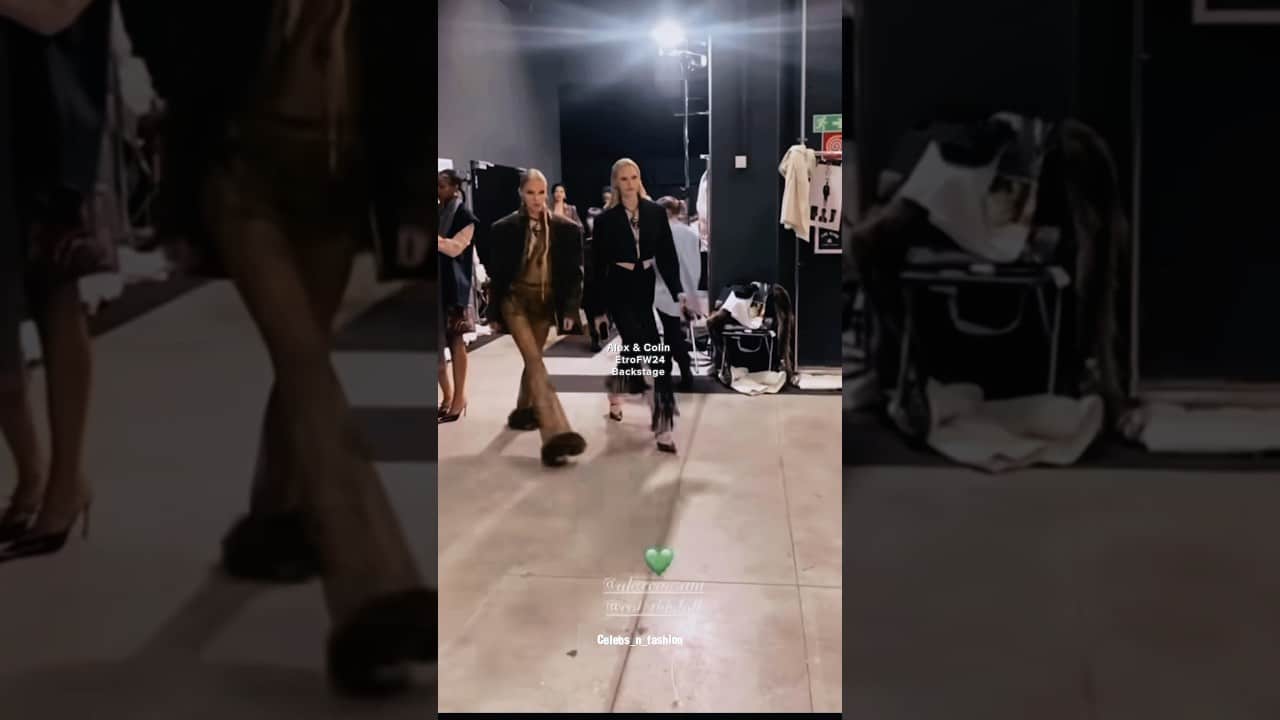 Models walking backstage at a fashion show, with clothing racks and scattered items in the background.