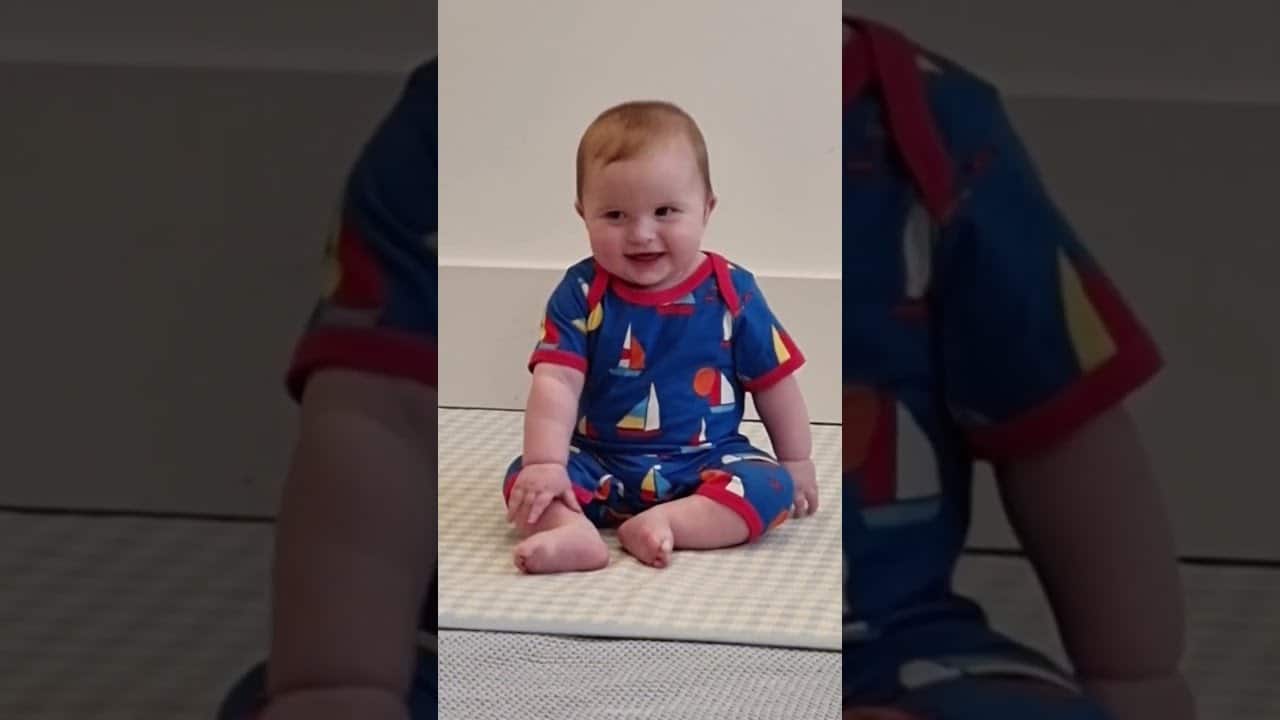 Baby wearing a blue onesie with colorful patterns sits on a padded mat against a plain background.